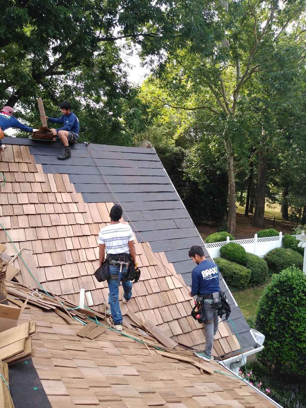 Two roofers on a steep shingle roof above a tree line